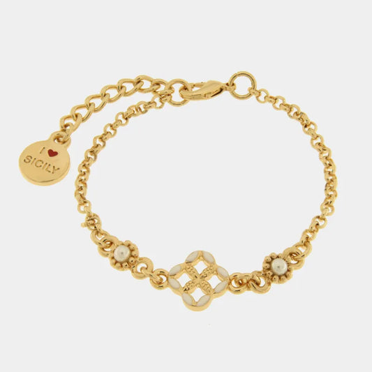 Bracciale in metallo con finitura dorata del marchio BySimon appartenente alla collezione Fiori di Lava caratterizzato da un design mediterraneo con maglia a catena rolo. Il gioiello presenta un elemento centrale a forma di quadrifoglio traforato ispirato alle maioliche siciliane, decorato con smalto bianco sui petali e un motivo a rilievo dorato nel mezzo. Ai lati del decoro centrale sono incastonate due piccole perle bianche circondate da una lavorazione granulata. Completano il bracciale una chiusura a 