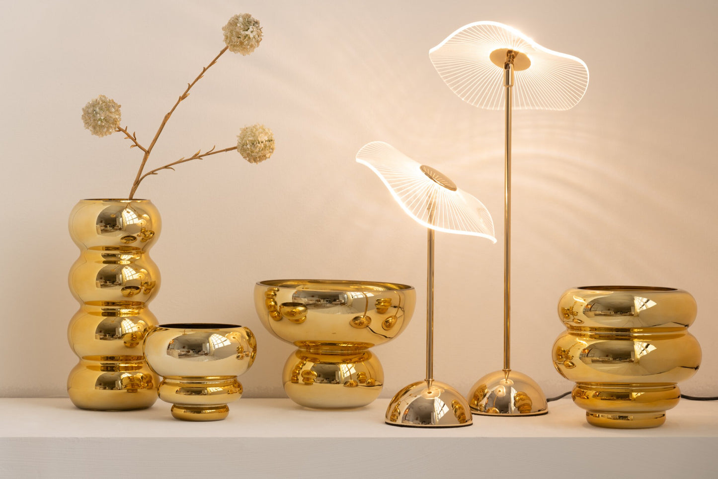 Set di portacandele e vasi decorativi assortiti della collezione Julia, abbinati a eleganti lampade da tavolo coordinate, tutti caratterizzati da una lussuosa finitura oro lucida e specchiata. Gli elementi presentano diverse forme bombate, sagomate con design a sfere e cerchi sovrapposti, ideali per creare composizioni scenografiche e infondere un'atmosfera calda, accogliente e di design negli ambienti interni.