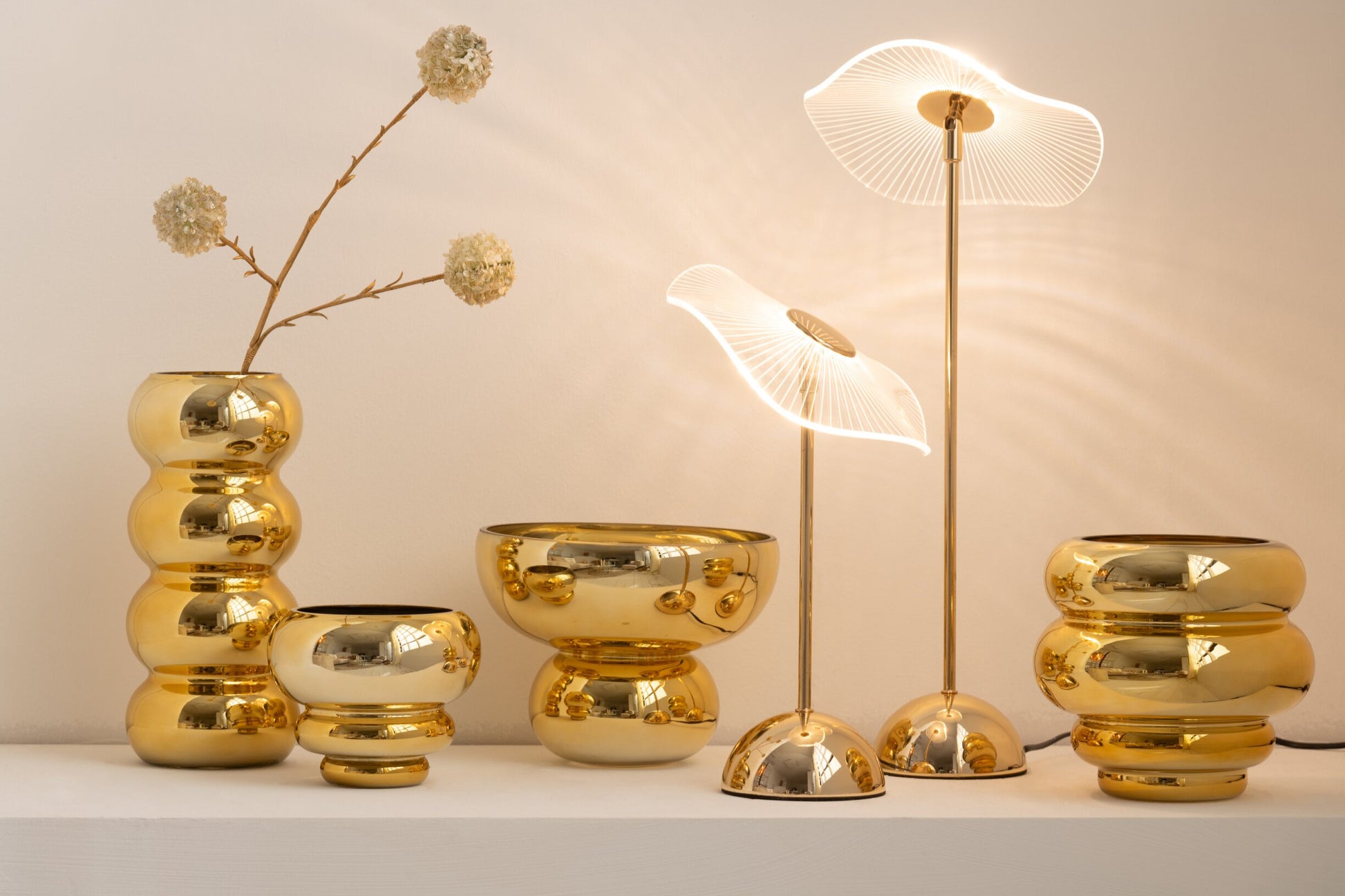 Set di portacandele e vasi decorativi assortiti della collezione Julia, abbinati a eleganti lampade da tavolo coordinate, tutti caratterizzati da una lussuosa finitura oro lucida e specchiata. Gli elementi presentano diverse forme bombate, sagomate con design a sfere e cerchi sovrapposti, ideali per creare composizioni scenografiche e infondere un'atmosfera calda, accogliente e di design negli ambienti interni.