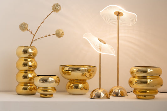 Set di portacandele e vasi decorativi assortiti della collezione Julia, abbinati a eleganti lampade da tavolo coordinate, tutti caratterizzati da una lussuosa finitura oro lucida e specchiata. Gli elementi presentano diverse forme bombate, sagomate con design a sfere e cerchi sovrapposti, ideali per creare composizioni scenografiche e infondere un'atmosfera calda, accogliente e di design negli ambienti interni.
