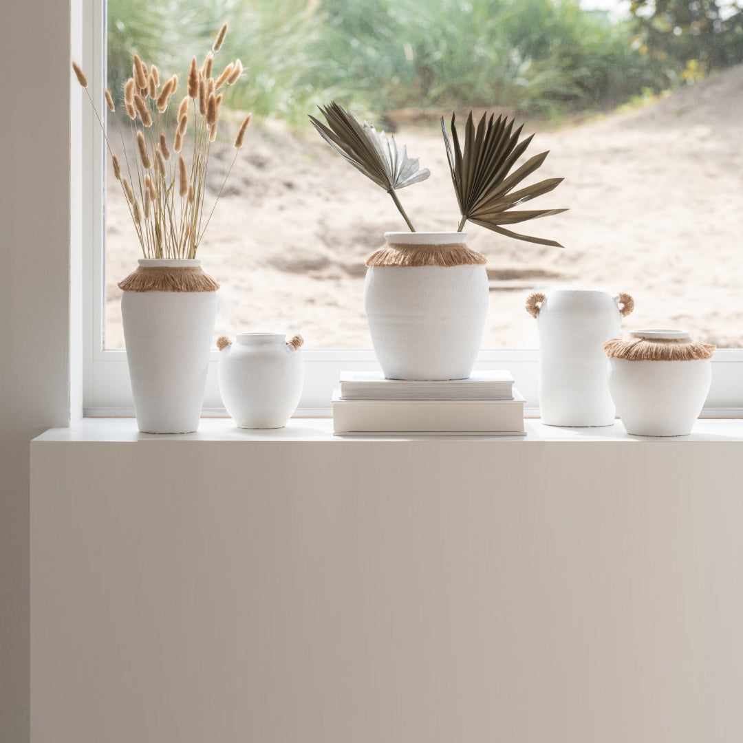 Vaso piccolo in ceramica della collezione Ibiza "Oceano Azul" di Jolipa, caratterizzato da una struttura bombata bianca con superficie dall'aspetto materico e naturale. Questo complemento d'arredo è decorato sulla parte superiore da un colletto in juta sfrangiata marrone, ideale per arricchire ambienti in stile boho chic o etnico.