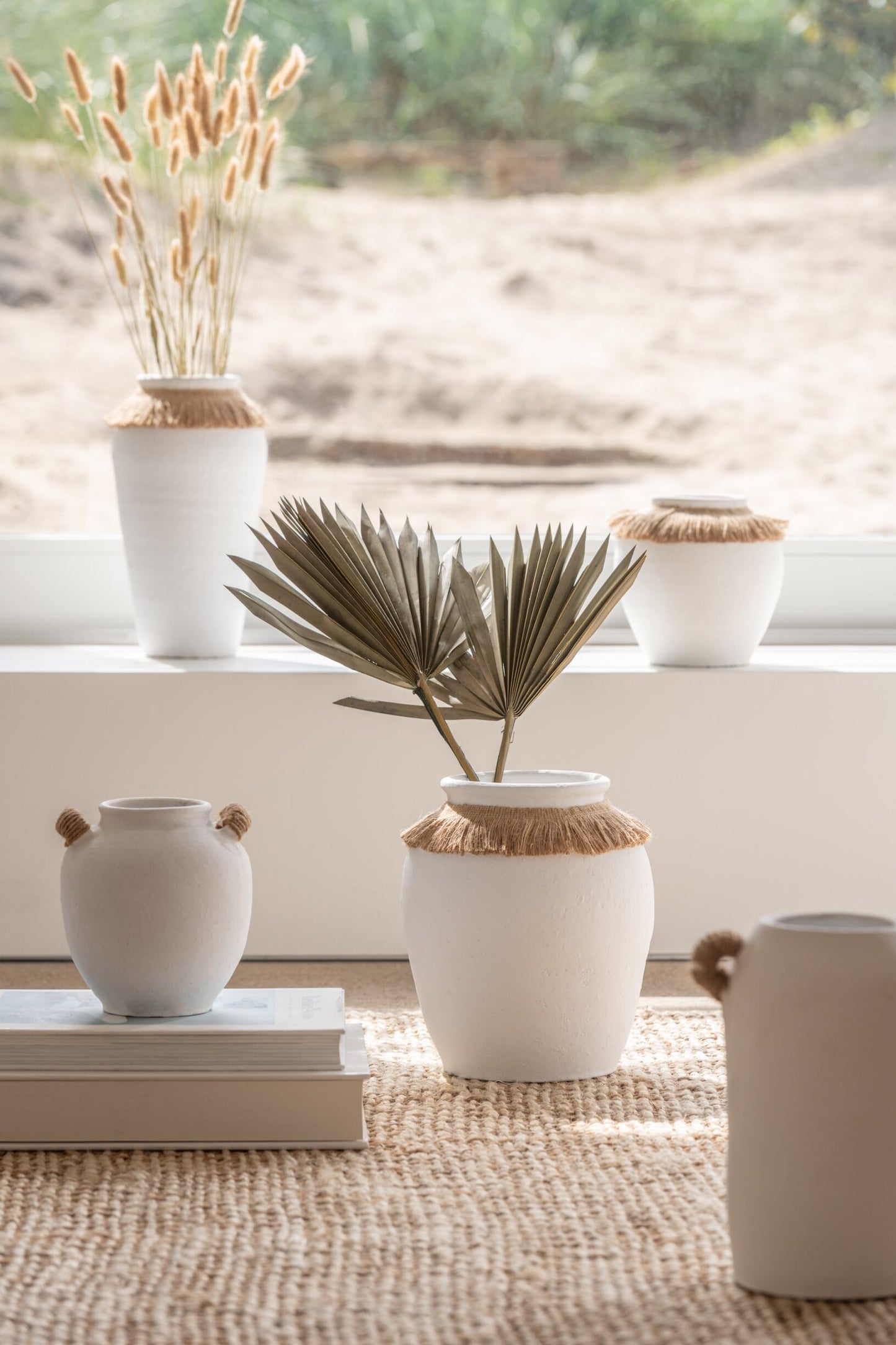 Vaso piccolo in ceramica della collezione Ibiza "Oceano Azul" di Jolipa, caratterizzato da una struttura bombata bianca con superficie dall'aspetto materico e naturale. Questo complemento d'arredo è decorato sulla parte superiore da un colletto in juta sfrangiata marrone, ideale per arricchire ambienti in stile boho chic o etnico.