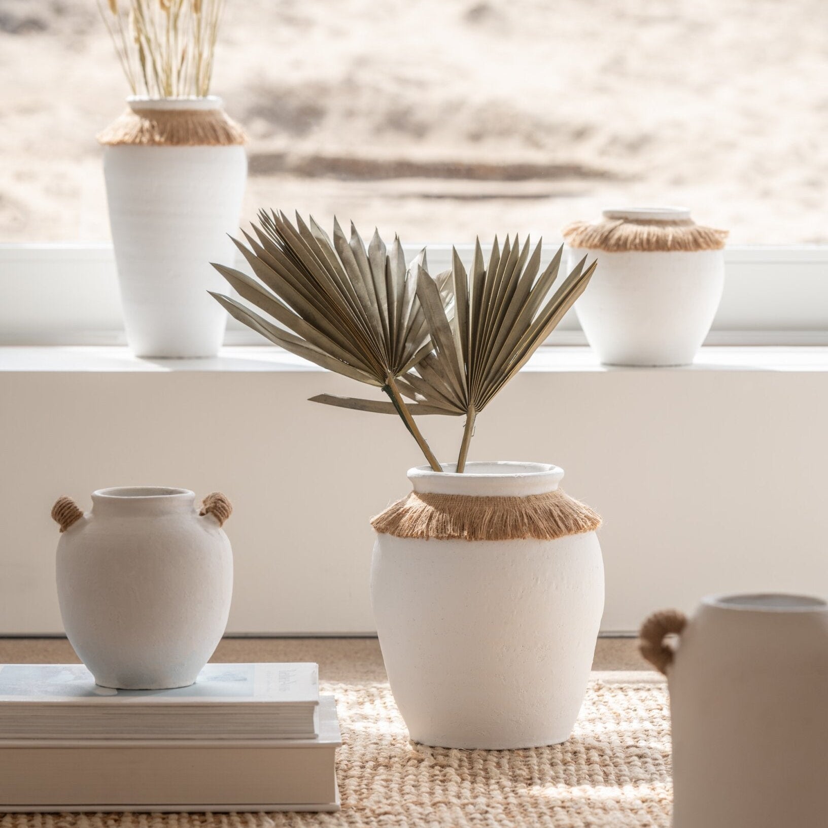 Vaso piccolo in ceramica della collezione Ibiza "Oceano Azul" di Jolipa, caratterizzato da una struttura bombata bianca con superficie dall'aspetto materico e naturale. Questo complemento d'arredo è decorato sulla parte superiore da un colletto in juta sfrangiata marrone, ideale per arricchire ambienti in stile boho chic o etnico.