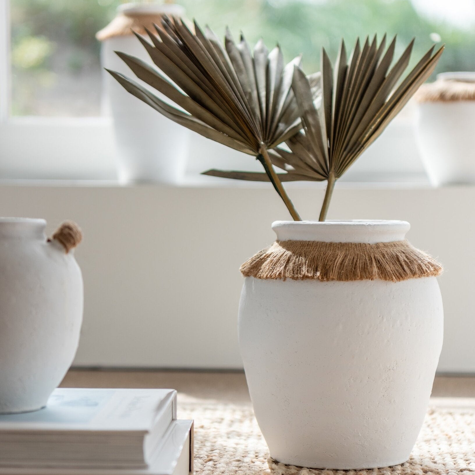 Vaso piccolo in ceramica della collezione Ibiza "Oceano Azul" di Jolipa, caratterizzato da una struttura bombata bianca con superficie dall'aspetto materico e naturale. Questo complemento d'arredo è decorato sulla parte superiore da un colletto in juta sfrangiata marrone, ideale per arricchire ambienti in stile boho chic o etnico.