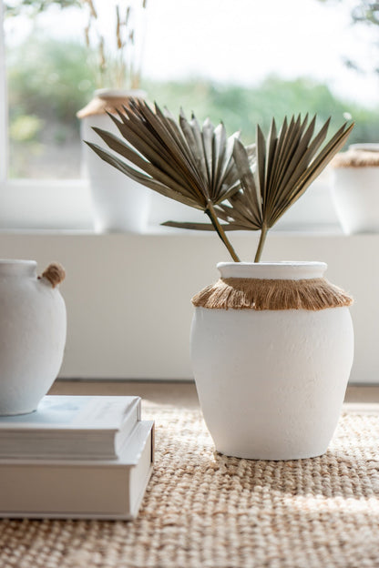 Vaso piccolo in ceramica della collezione Ibiza "Oceano Azul" di Jolipa, caratterizzato da una struttura bombata bianca con superficie dall'aspetto materico e naturale. Questo complemento d'arredo è decorato sulla parte superiore da un colletto in juta sfrangiata marrone, ideale per arricchire ambienti in stile boho chic o etnico.