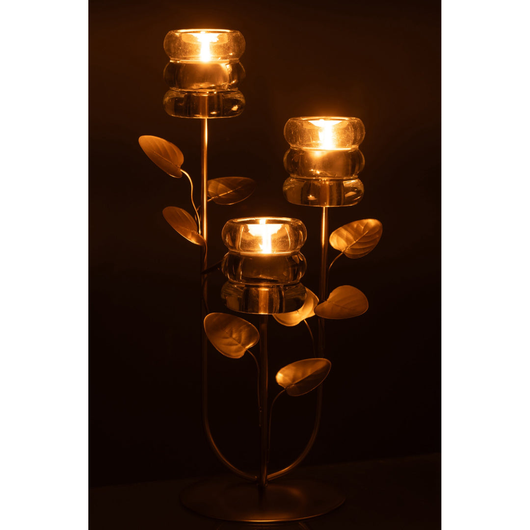 Candeliere decorativo a tre bracci caratterizzato da una struttura metallica dorata con steli disposti a rami ricchi di foglie decorative, che si innalzano da una base circolare satinata. Ognuna delle tre estremità supporta un elegante portacandele in vetro trasparente spesso con lavorazione a rilievo a cerchi sovrapposti, ideale come raffinato centrotavola per creare un'atmosfera calda e accogliente nei tuoi ambienti.