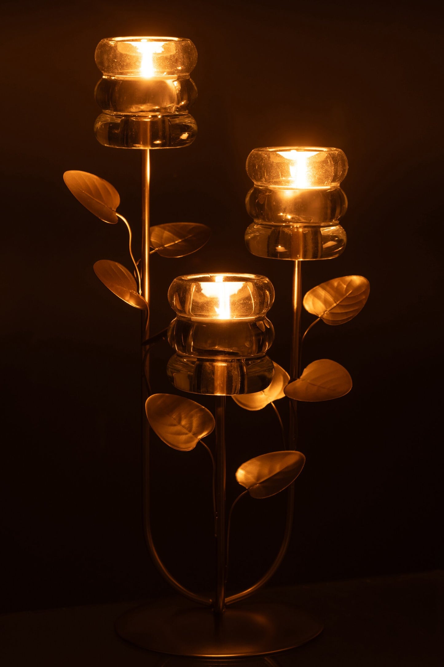 Candeliere decorativo a tre bracci caratterizzato da una struttura metallica dorata con steli disposti a rami ricchi di foglie decorative, che si innalzano da una base circolare satinata. Ognuna delle tre estremità supporta un elegante portacandele in vetro trasparente spesso con lavorazione a rilievo a cerchi sovrapposti, ideale come raffinato centrotavola per creare un'atmosfera calda e accogliente nei tuoi ambienti.