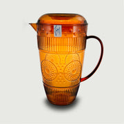 Una caraffa in plastica dal colore ambra trasparente, caratterizzata da una raffinata lavorazione a rilievo floreale e righe verticali che ne movimentano la superficie, richiamando lo stile del cristallo lavorato. Dotata di beccuccio versatore, coperchio e comoda manico ad ansa, unisce estetica vintage e praticità d'uso. Un accessorio dal gusto elegante e colorato, perfetto per la tavola estiva o per servire bevande fresche con stile.