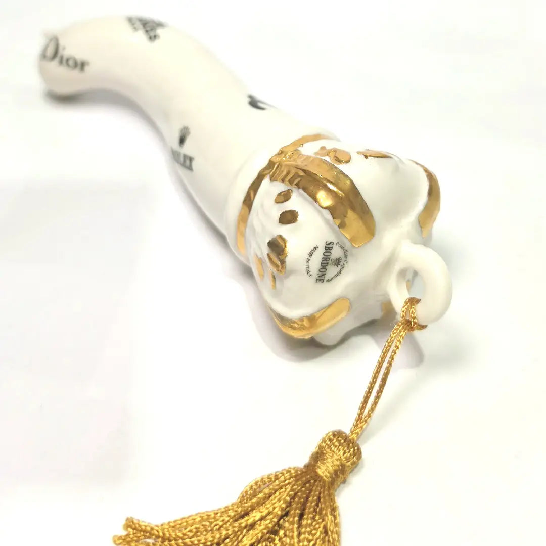 Corno portafortuna in ceramica bianca lucida decorato con loghi fashion neri e completato da una corona regale dorata con nappa in seta oro. Questo elegante oggetto scaramantico di design artigianale rappresenta un complemento d'arredo pop lussuoso e originale, perfetto come idea regalo o decorazione d'interni moderna.