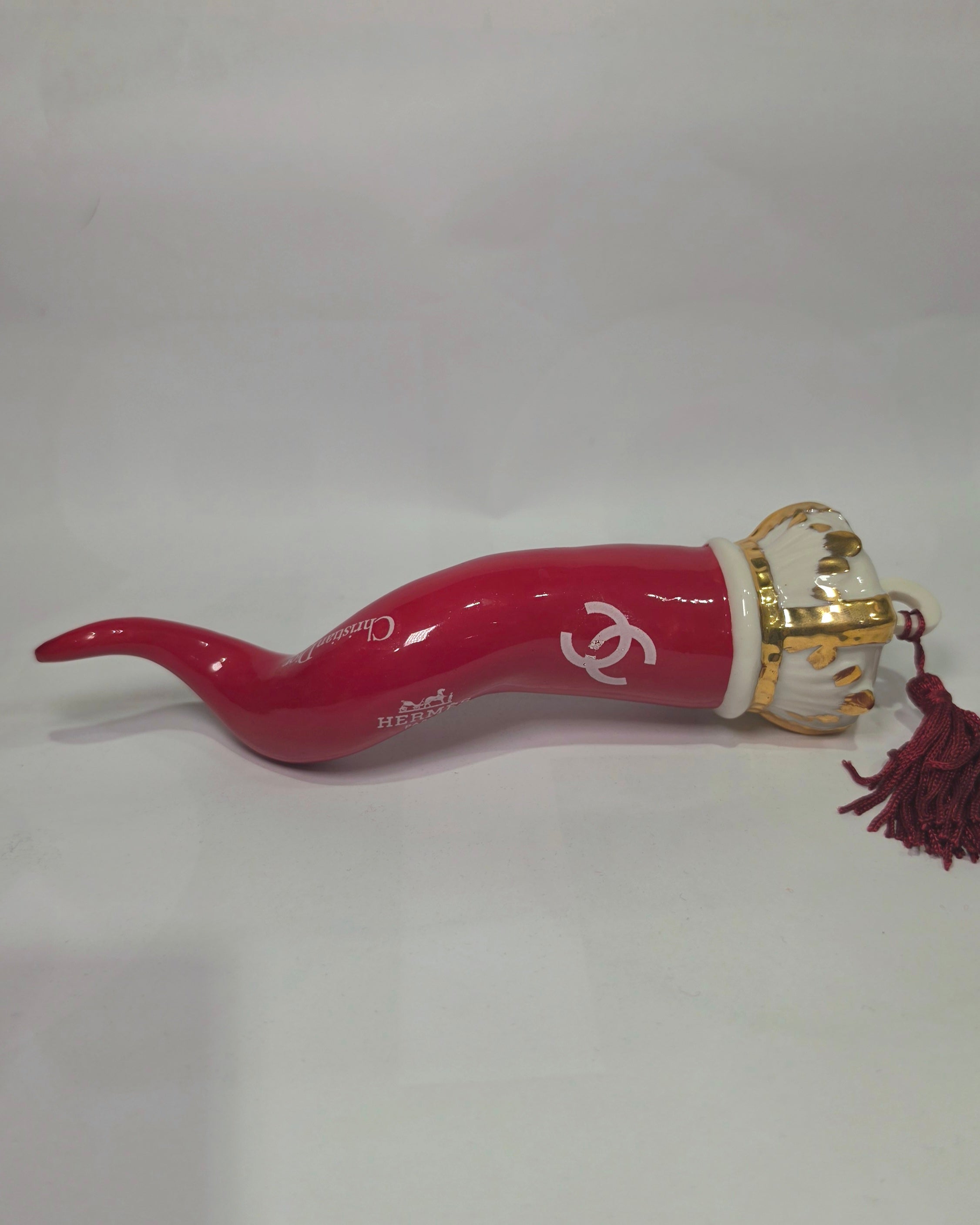 Corno portafortuna in ceramica rossa lucida decorato con loghi fashion bianchi e completato da una corona regale dorata con nappa in seta rossa. Questo elegante oggetto scaramantico di design artigianale rappresenta un complemento d'arredo pop lussuoso e originale, perfetto come idea regalo o decorazione d'interni moderna.
