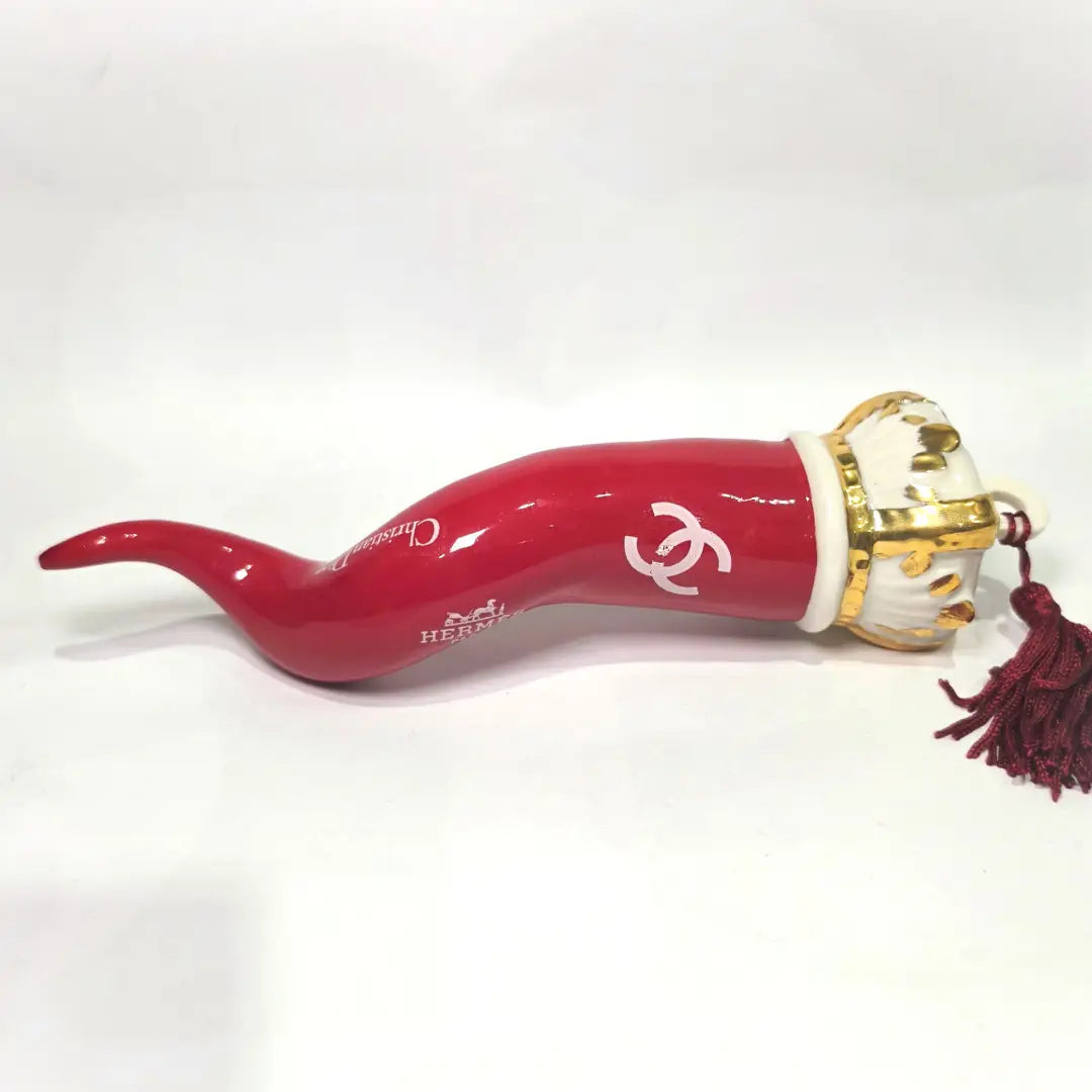 Corno portafortuna in ceramica rossa lucida decorato con loghi fashion bianchi e completato da una corona regale dorata con nappa in seta rossa. Questo elegante oggetto scaramantico di design artigianale rappresenta un complemento d'arredo pop lussuoso e originale, perfetto come idea regalo o decorazione d'interni moderna.