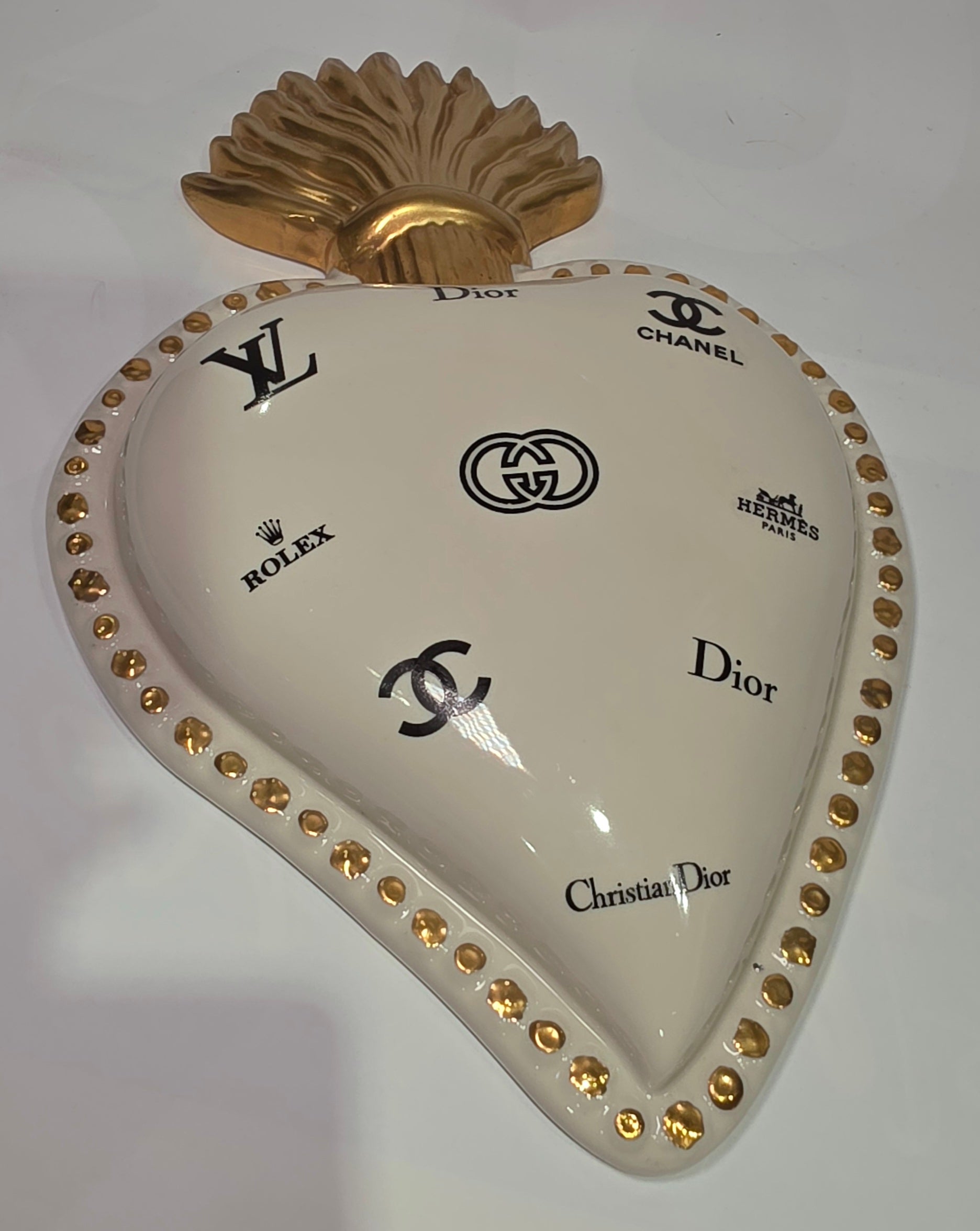 Scultura da parete a forma di Cuore Sacro grande in ceramica bianca lucida, decorata con molteplici loghi fashion neri e sormontata da una fiamma dorata finemente lavorata. L'oggetto presenta una cornice perlinata con borchie oro ed è un complemento d'arredo pop art di grande impatto visivo, ideale per personalizzare pareti in ambienti moderni e di lusso.