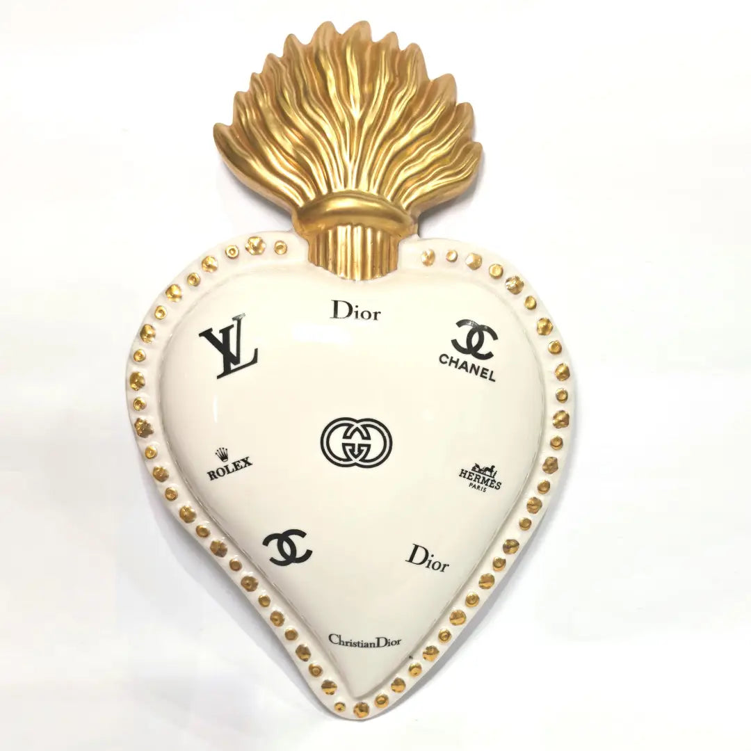 Scultura da parete a forma di Cuore Sacro grande in ceramica bianca lucida, decorata con molteplici loghi fashion neri e sormontata da una fiamma dorata finemente lavorata. L'oggetto presenta una cornice perlinata con borchie oro ed è un complemento d'arredo pop art di grande impatto visivo, ideale per personalizzare pareti in ambienti moderni e di lusso.