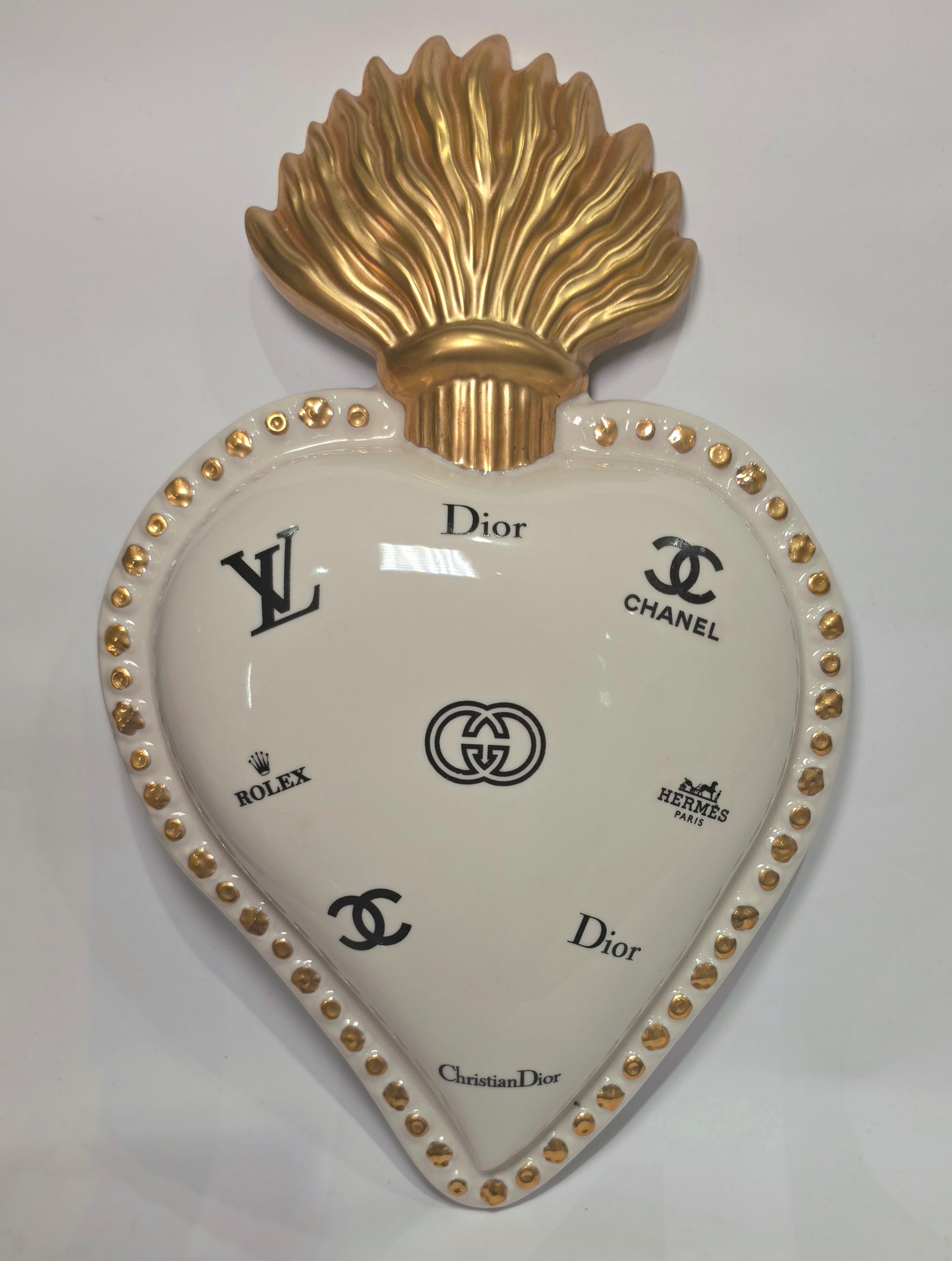 Scultura da parete a forma di Cuore Sacro grande in ceramica bianca lucida, decorata con molteplici loghi fashion neri e sormontata da una fiamma dorata finemente lavorata. L'oggetto presenta una cornice perlinata con borchie oro ed è un complemento d'arredo pop art di grande impatto visivo, ideale per personalizzare pareti in ambienti moderni e di lusso.