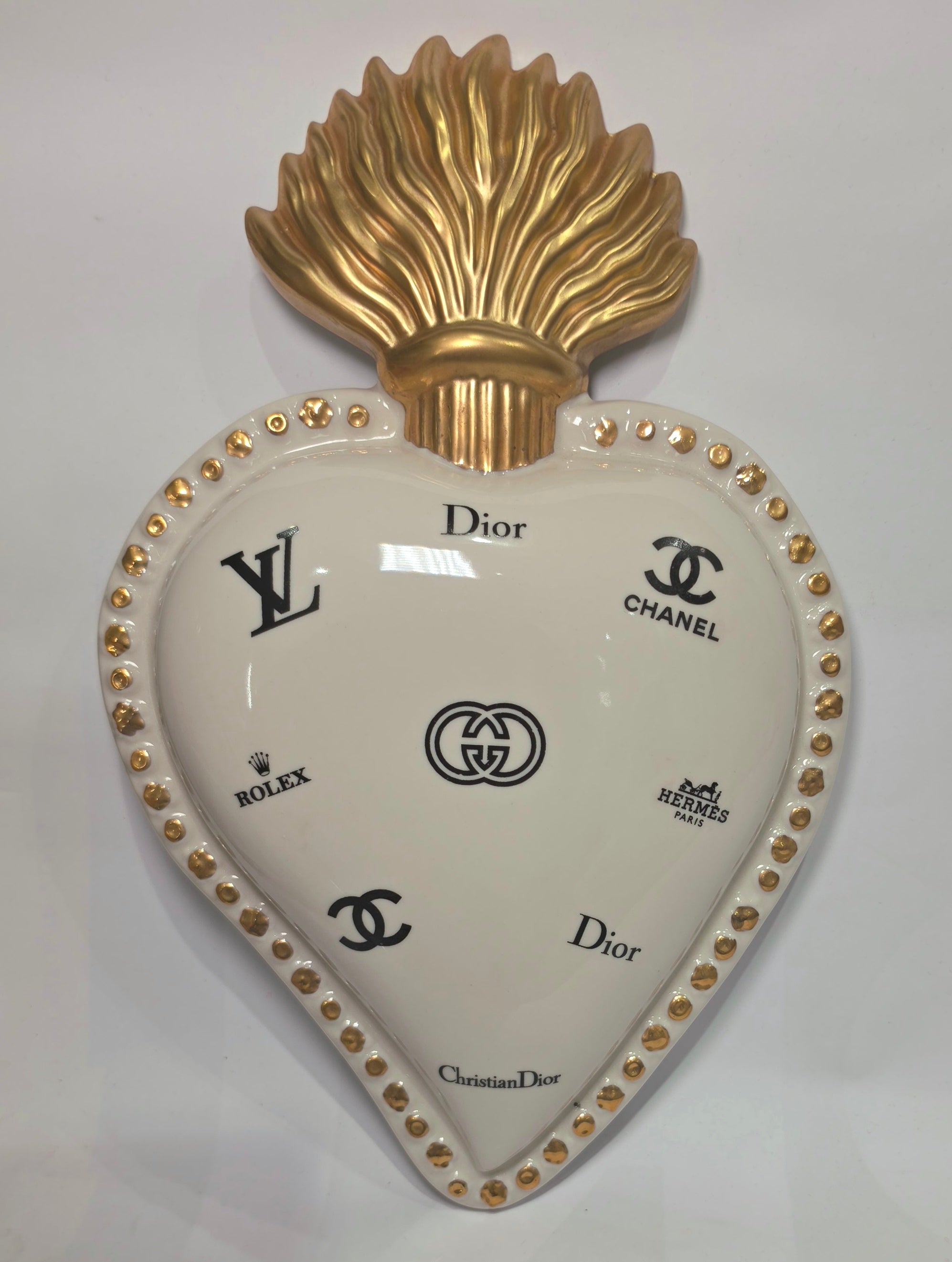 Scultura da parete a forma di Cuore Sacro grande in ceramica bianca lucida, decorata con molteplici loghi fashion neri e sormontata da una fiamma dorata finemente lavorata. L'oggetto presenta una cornice perlinata con borchie oro ed è un complemento d'arredo pop art di grande impatto visivo, ideale per personalizzare pareti in ambienti moderni e di lusso.