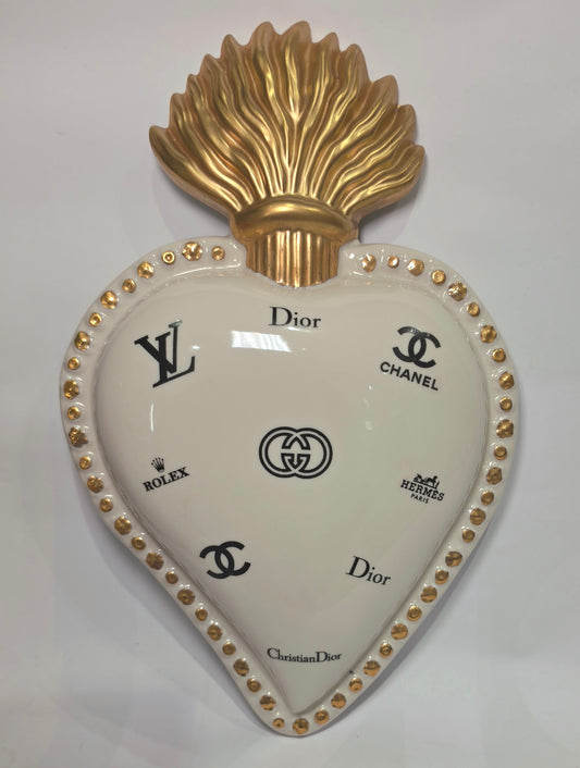 Scultura da parete a forma di Cuore Sacro grande in ceramica bianca lucida, decorata con molteplici loghi fashion neri e sormontata da una fiamma dorata finemente lavorata. L'oggetto presenta una cornice perlinata con borchie oro ed è un complemento d'arredo pop art di grande impatto visivo, ideale per personalizzare pareti in ambienti moderni e di lusso.