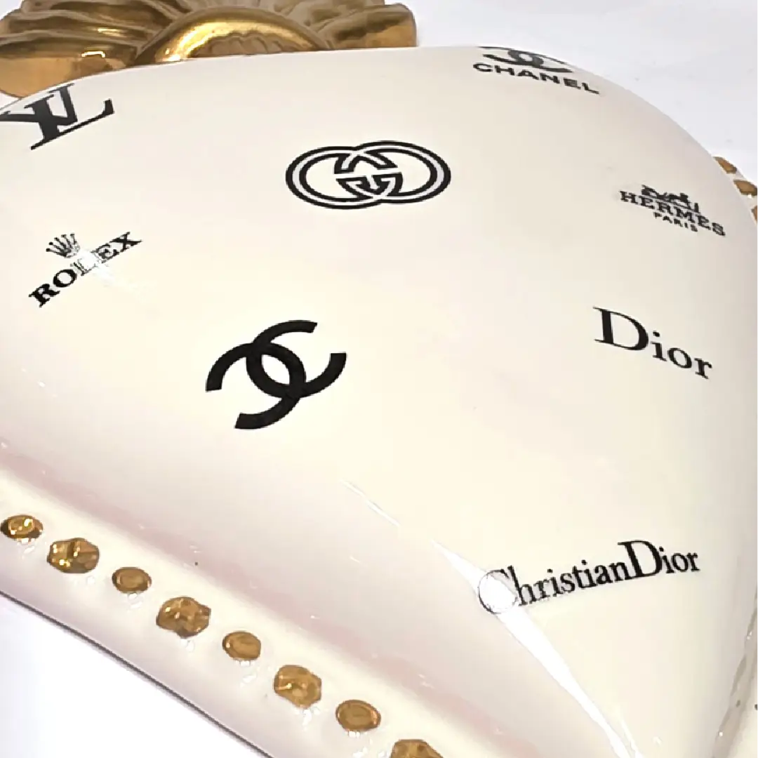 Scultura da parete a forma di Cuore Sacro grande in ceramica bianca lucida, decorata con molteplici loghi fashion neri e sormontata da una fiamma dorata finemente lavorata. L'oggetto presenta una cornice perlinata con borchie oro ed è un complemento d'arredo pop art di grande impatto visivo, ideale per personalizzare pareti in ambienti moderni e di lusso.