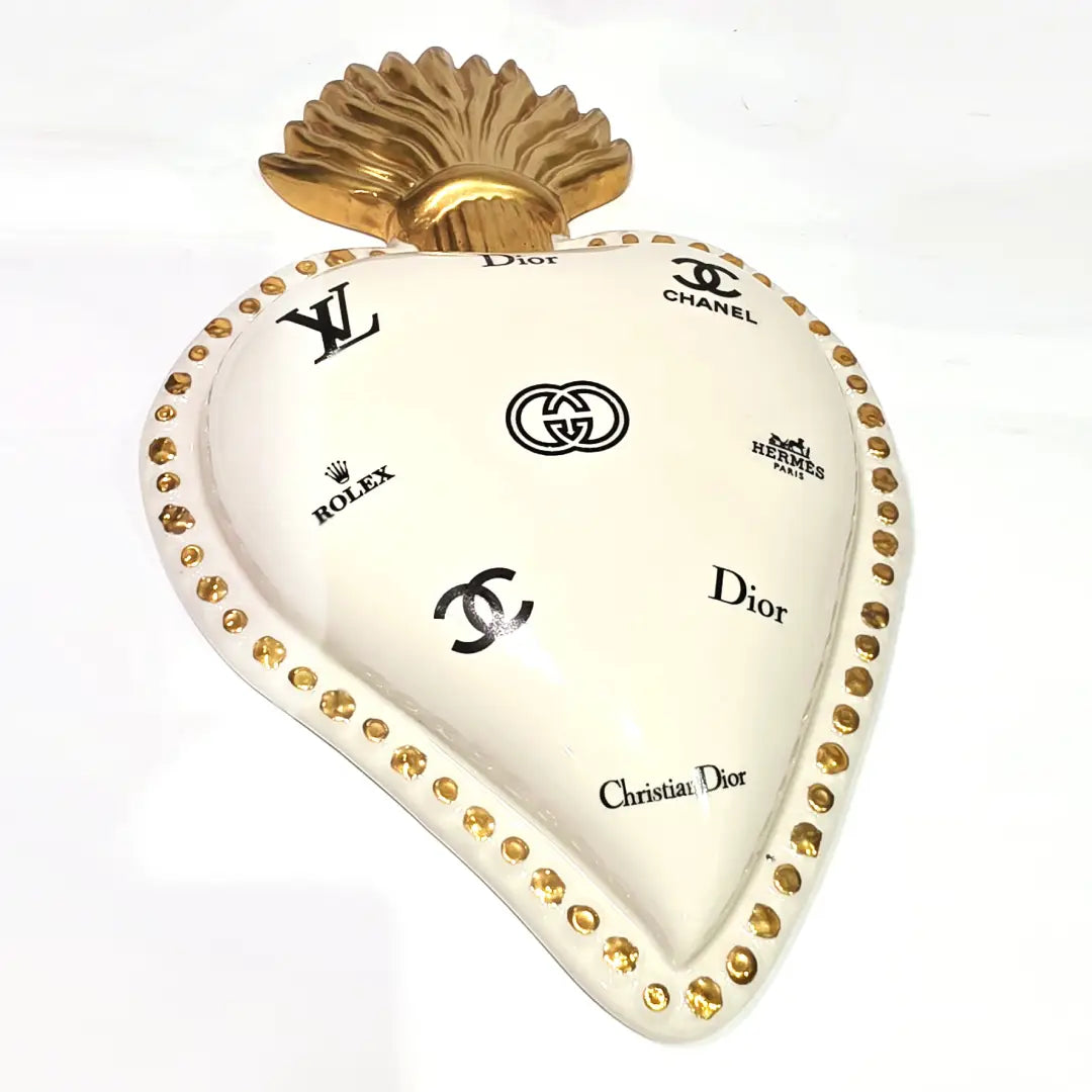 Scultura da parete a forma di Cuore Sacro grande in ceramica bianca lucida, decorata con molteplici loghi fashion neri e sormontata da una fiamma dorata finemente lavorata. L'oggetto presenta una cornice perlinata con borchie oro ed è un complemento d'arredo pop art di grande impatto visivo, ideale per personalizzare pareti in ambienti moderni e di lusso.