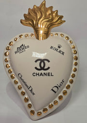 Scultura decorativa a forma di Cuore Sacro in ceramica bianca lucida, impreziosita da loghi fashion neri a contrasto e una maestosa fiamma superiore dorata. Questo complemento d'arredo di design presenta una cornice perlinata con borchie oro e una base d'appoggio, ideale per decorare ambienti moderni con un tocco pop art esclusivo.