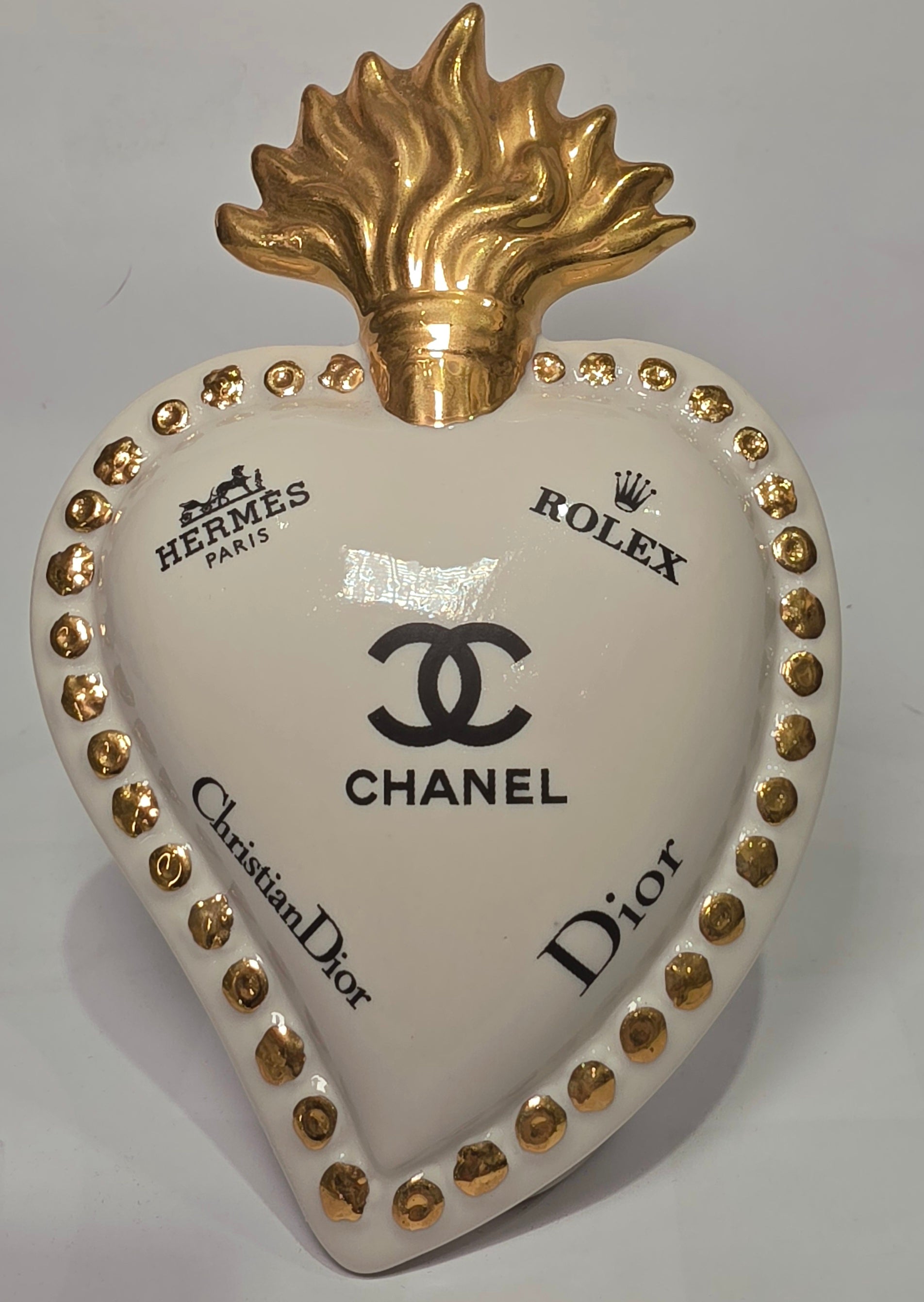 Scultura decorativa a forma di Cuore Sacro in ceramica bianca lucida, impreziosita da loghi fashion neri a contrasto e una maestosa fiamma superiore dorata. Questo complemento d'arredo di design presenta una cornice perlinata con borchie oro e una base d'appoggio, ideale per decorare ambienti moderni con un tocco pop art esclusivo.