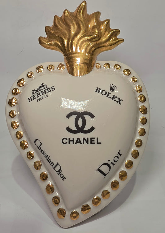Scultura decorativa a forma di Cuore Sacro in ceramica bianca lucida, impreziosita da loghi fashion neri a contrasto e una maestosa fiamma superiore dorata. Questo complemento d'arredo di design presenta una cornice perlinata con borchie oro e una base d'appoggio, ideale per decorare ambienti moderni con un tocco pop art esclusivo.