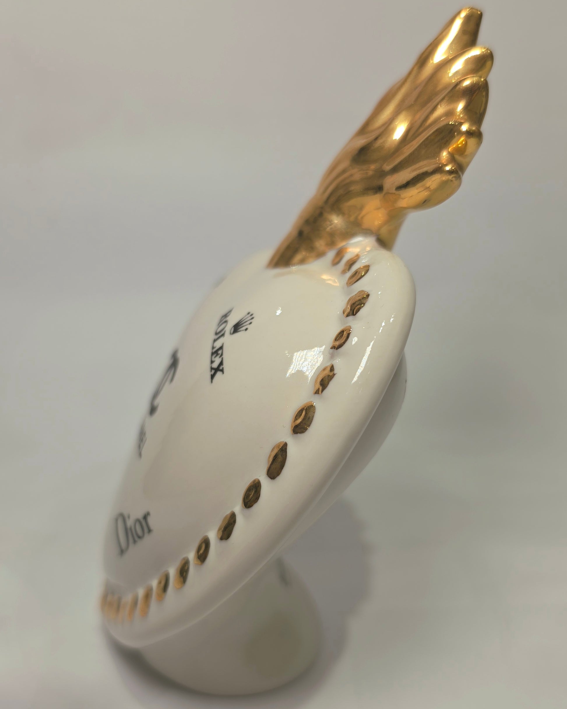 Scultura decorativa a forma di Cuore Sacro in ceramica bianca lucida, impreziosita da loghi fashion neri a contrasto e una maestosa fiamma superiore dorata. Questo complemento d'arredo di design presenta una cornice perlinata con borchie oro e una base d'appoggio, ideale per decorare ambienti moderni con un tocco pop art esclusivo.
