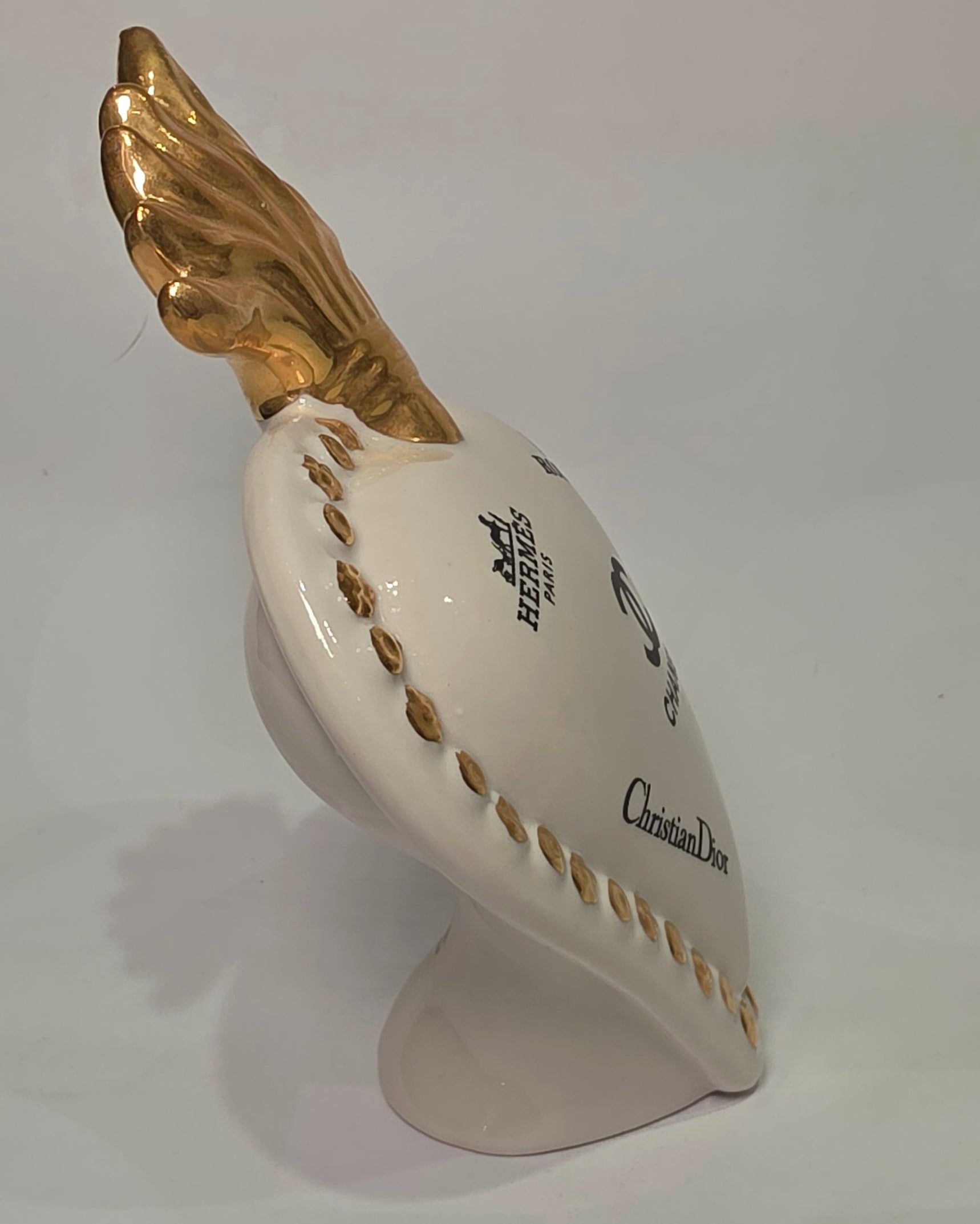 Scultura decorativa a forma di Cuore Sacro in ceramica bianca lucida, impreziosita da loghi fashion neri a contrasto e una maestosa fiamma superiore dorata. Questo complemento d'arredo di design presenta una cornice perlinata con borchie oro e una base d'appoggio, ideale per decorare ambienti moderni con un tocco pop art esclusivo.
