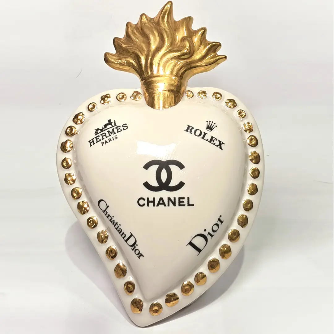 Scultura decorativa a forma di Cuore Sacro in ceramica bianca lucida, impreziosita da loghi fashion neri a contrasto e una maestosa fiamma superiore dorata. Questo complemento d'arredo di design presenta una cornice perlinata con borchie oro e una base d'appoggio, ideale per decorare ambienti moderni con un tocco pop art esclusivo.