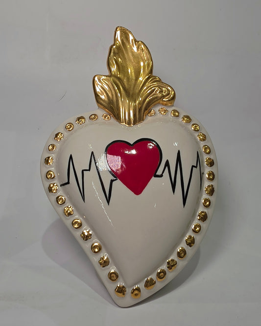 Scultura decorativa a forma di Cuore Sacro in ceramica bianca lucida, caratterizzata da un cuore rosso centrale sovrapposto a un disegno grafico nero che richiama il battito cardiaco. L'oggetto è impreziosito da una fiamma superiore dorata e una cornice perlinata con borchie oro, rendendolo un complemento d'arredo dal design moderno e originale.