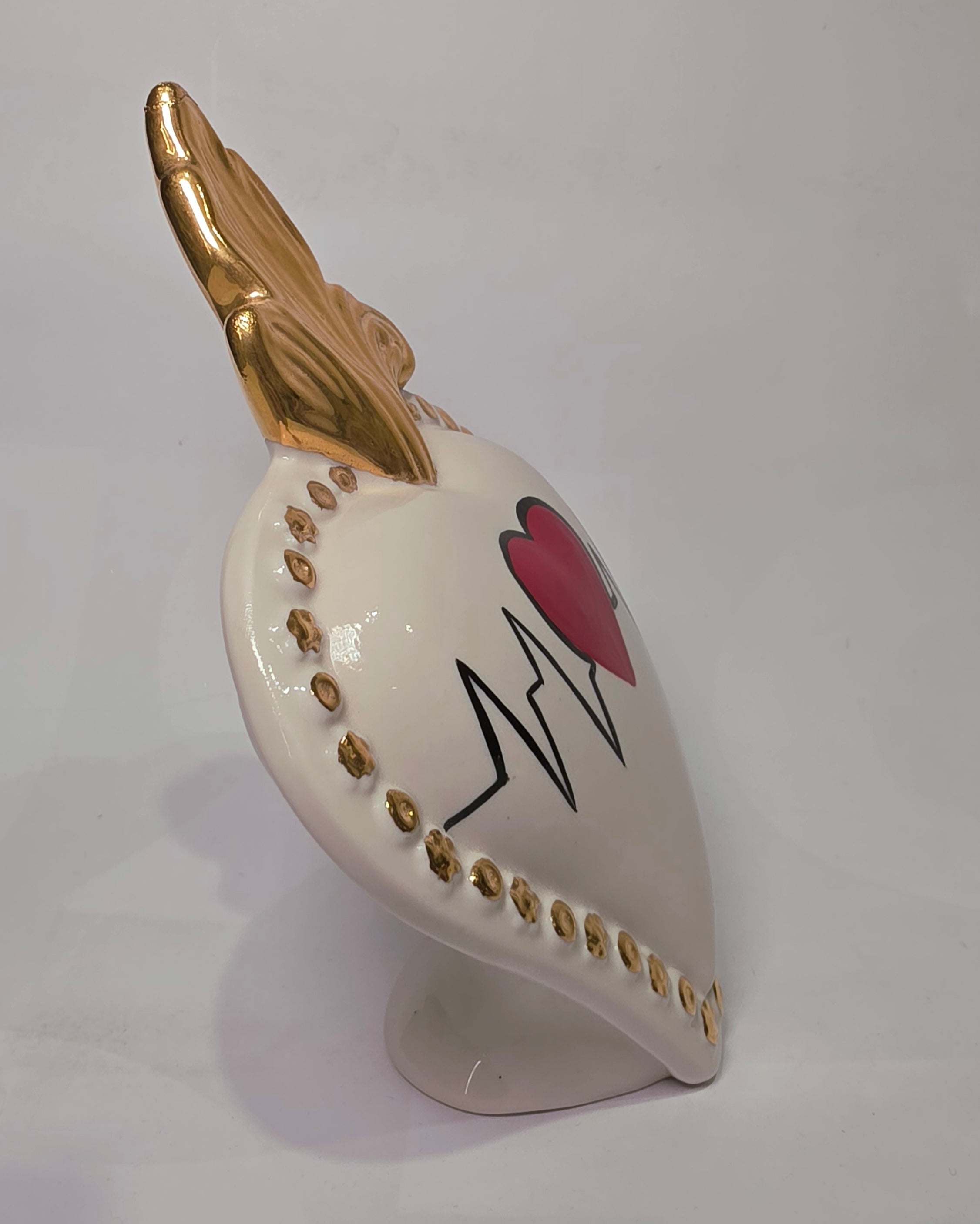 Scultura decorativa a forma di Cuore Sacro in ceramica bianca lucida, caratterizzata da un cuore rosso centrale sovrapposto a un disegno grafico nero che richiama il battito cardiaco. L'oggetto è impreziosito da una fiamma superiore dorata e una cornice perlinata con borchie oro, rendendolo un complemento d'arredo dal design moderno e originale.
