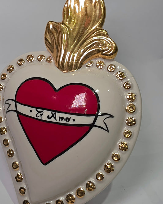 Scultura decorativa a forma di Cuore Sacro in ceramica bianca lucida, decorata al centro con un cuore rosso e una pergamena con la scritta "Ti Amo". L'oggetto è impreziosito da una fiamma superiore dorata e una cornice perlinata con borchie oro, rappresentando un'idea regalo romantica e un complemento d'arredo dal design unico.