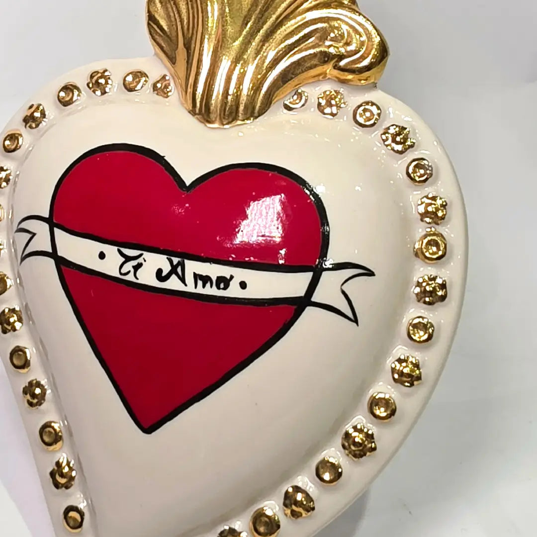 Scultura decorativa a forma di Cuore Sacro in ceramica bianca lucida, decorata al centro con un cuore rosso e una pergamena con la scritta "Ti Amo". L'oggetto è impreziosito da una fiamma superiore dorata e una cornice perlinata con borchie oro, rappresentando un'idea regalo romantica e un complemento d'arredo dal design unico.