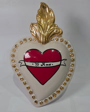 Scultura decorativa a forma di Cuore Sacro in ceramica bianca lucida, decorata al centro con un cuore rosso e una pergamena con la scritta "Ti Amo". L'oggetto è impreziosito da una fiamma superiore dorata e una cornice perlinata con borchie oro, rappresentando un'idea regalo romantica e un complemento d'arredo dal design unico.