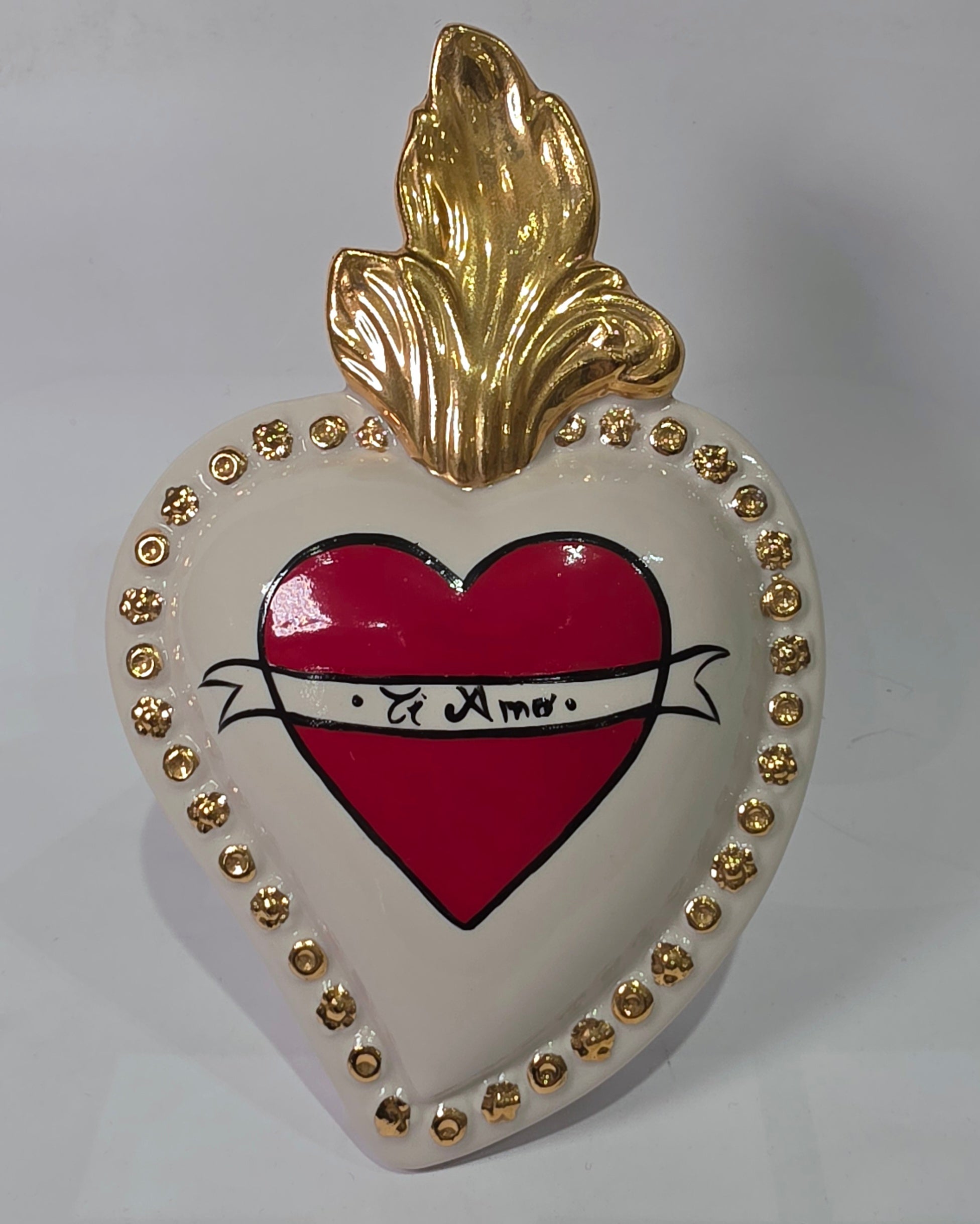 Scultura decorativa a forma di Cuore Sacro in ceramica bianca lucida, decorata al centro con un cuore rosso e una pergamena con la scritta "Ti Amo". L'oggetto è impreziosito da una fiamma superiore dorata e una cornice perlinata con borchie oro, rappresentando un'idea regalo romantica e un complemento d'arredo dal design unico.