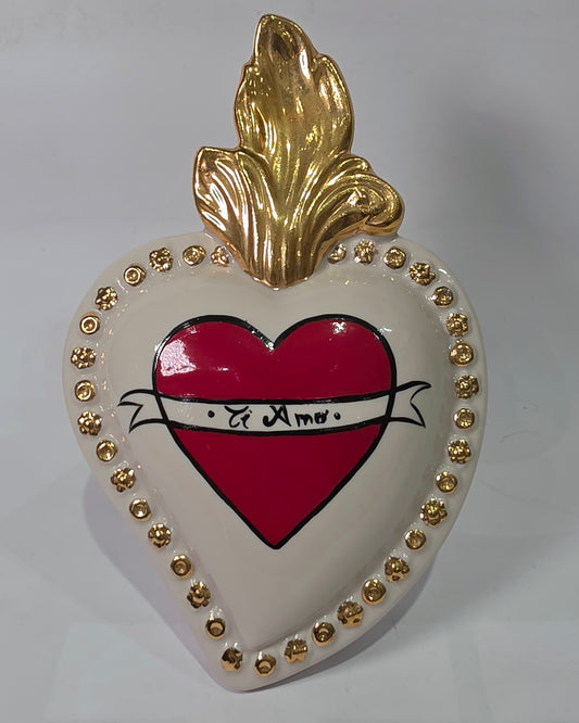 Scultura decorativa a forma di Cuore Sacro in ceramica bianca lucida, decorata al centro con un cuore rosso e una pergamena con la scritta "Ti Amo". L'oggetto è impreziosito da una fiamma superiore dorata e una cornice perlinata con borchie oro, rappresentando un'idea regalo romantica e un complemento d'arredo dal design unico.