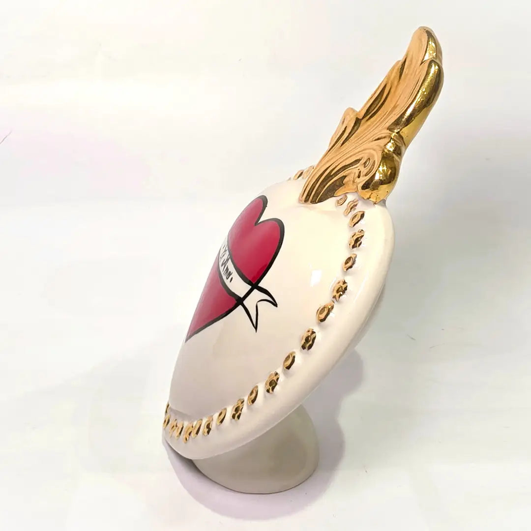 Scultura decorativa a forma di Cuore Sacro in ceramica bianca lucida, decorata al centro con un cuore rosso e una pergamena con la scritta "Ti Amo". L'oggetto è impreziosito da una fiamma superiore dorata e una cornice perlinata con borchie oro, rappresentando un'idea regalo romantica e un complemento d'arredo dal design unico.