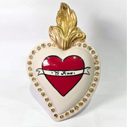 Scultura decorativa a forma di Cuore Sacro in ceramica bianca lucida, decorata al centro con un cuore rosso e una pergamena con la scritta "Ti Amo". L'oggetto è impreziosito da una fiamma superiore dorata e una cornice perlinata con borchie oro, rappresentando un'idea regalo romantica e un complemento d'arredo dal design unico.