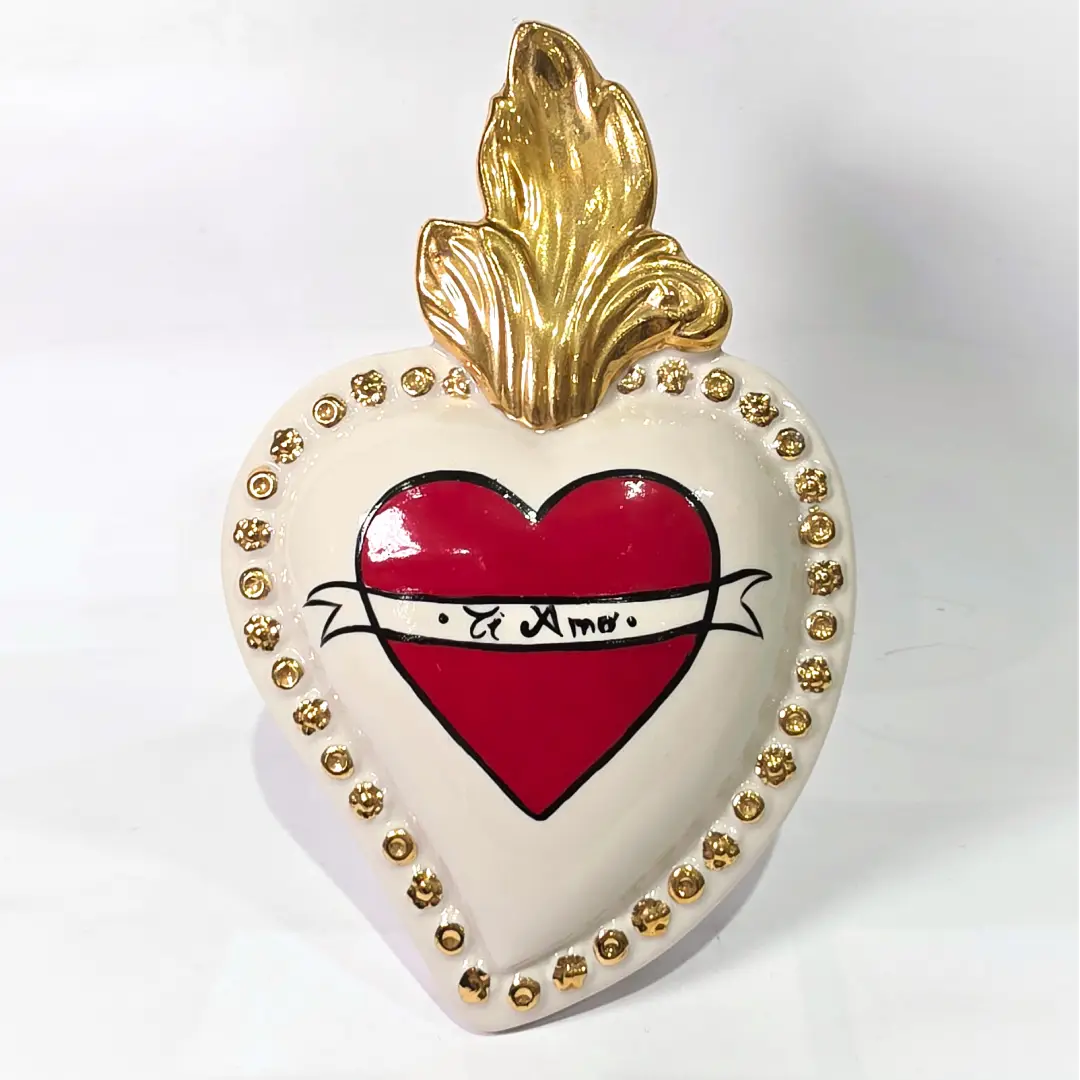 Scultura decorativa a forma di Cuore Sacro in ceramica bianca lucida, decorata al centro con un cuore rosso e una pergamena con la scritta "Ti Amo". L'oggetto è impreziosito da una fiamma superiore dorata e una cornice perlinata con borchie oro, rappresentando un'idea regalo romantica e un complemento d'arredo dal design unico.
