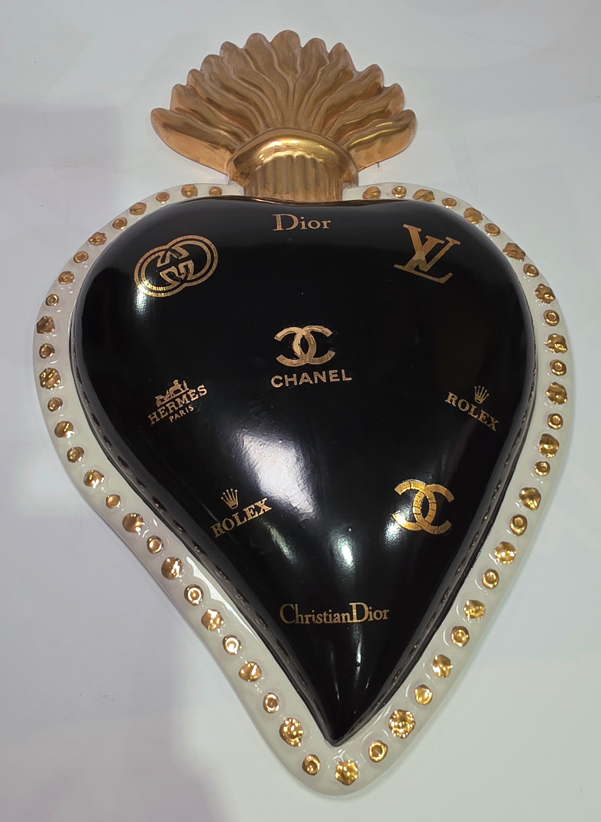 Scultura da parete a forma di Cuore Sacro grande in ceramica nera lucida, impreziosita da numerosi loghi fashion dorati e sormontata da una fiamma in oro finemente scolpita. Questo complemento d'arredo pop art presenta una cornice bianca perlinata con borchie oro, perfetto per decorare con lusso e originalità gli interni di una casa moderna.