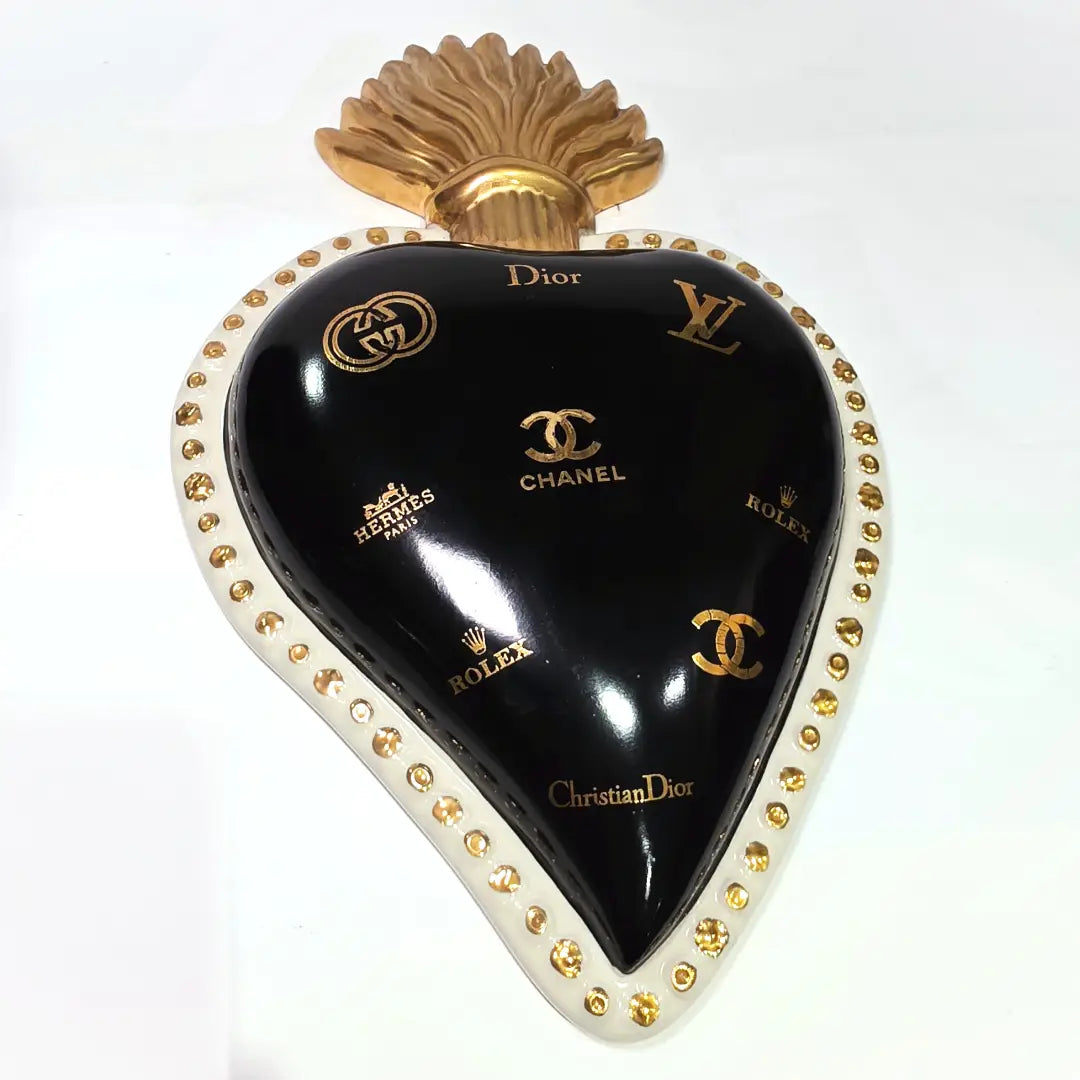 Scultura da parete a forma di Cuore Sacro grande in ceramica nera lucida, impreziosita da numerosi loghi fashion dorati e sormontata da una fiamma in oro finemente scolpita. Questo complemento d'arredo pop art presenta una cornice bianca perlinata con borchie oro, perfetto per decorare con lusso e originalità gli interni di una casa moderna.