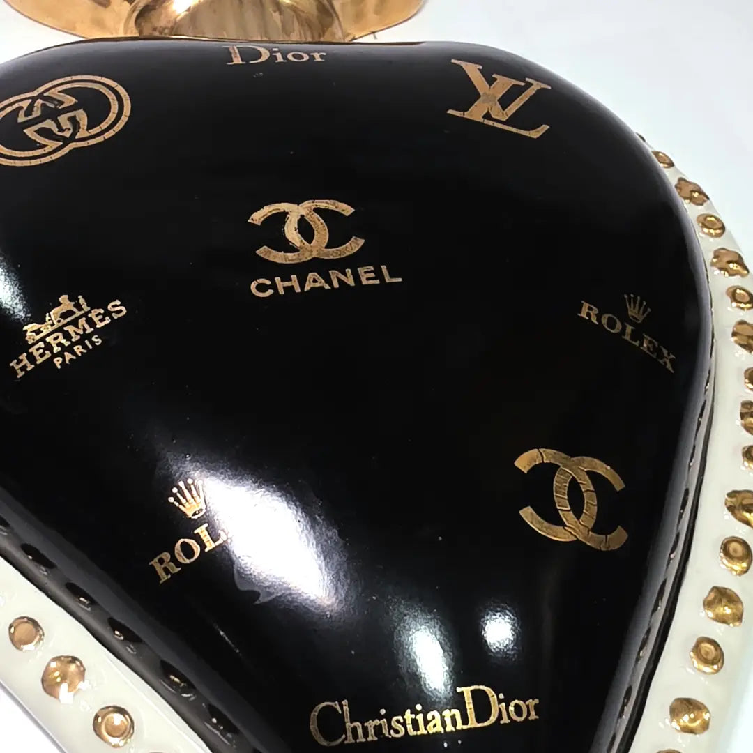 Scultura da parete a forma di Cuore Sacro grande in ceramica nera lucida, impreziosita da numerosi loghi fashion dorati e sormontata da una fiamma in oro finemente scolpita. Questo complemento d'arredo pop art presenta una cornice bianca perlinata con borchie oro, perfetto per decorare con lusso e originalità gli interni di una casa moderna.