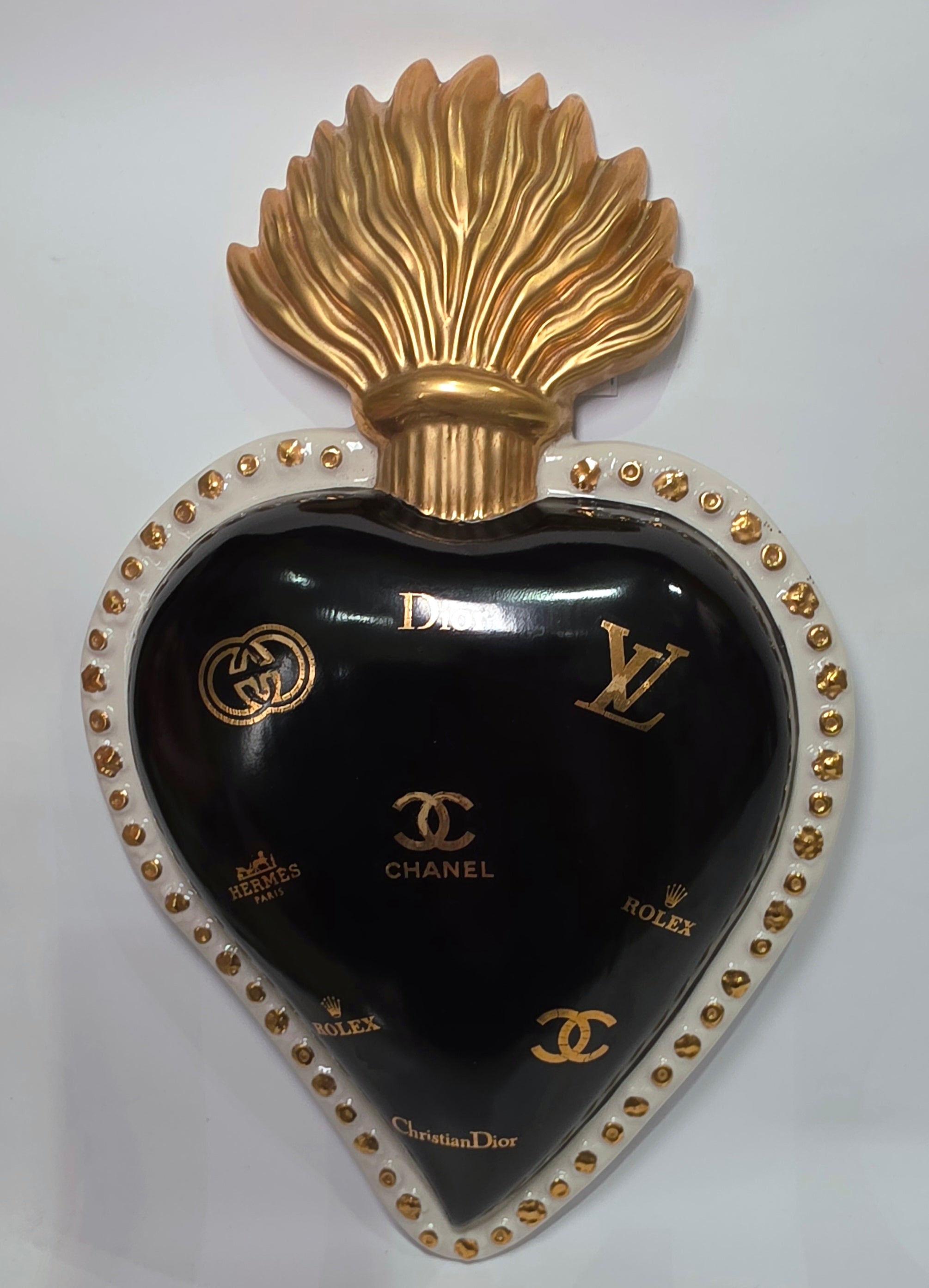 Scultura da parete a forma di Cuore Sacro grande in ceramica nera lucida, impreziosita da numerosi loghi fashion dorati e sormontata da una fiamma in oro finemente scolpita. Questo complemento d'arredo pop art presenta una cornice bianca perlinata con borchie oro, perfetto per decorare con lusso e originalità gli interni di una casa moderna.
