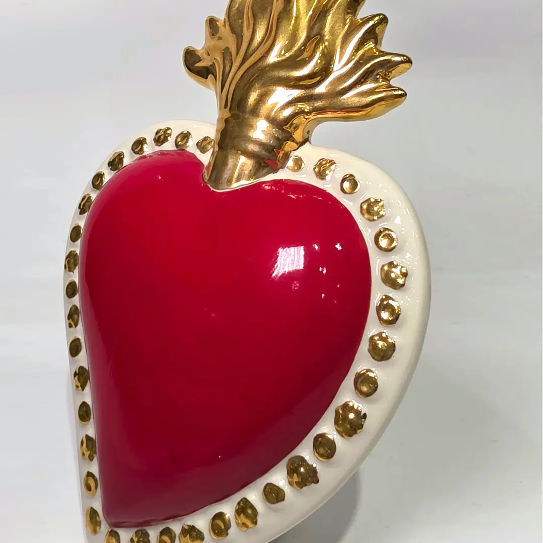 Scultura a forma di Cuore Sacro in ceramica rossa lucida, completata da una fiamma superiore dorata e una cornice bianca perlinata con borchie oro. Questo oggetto di design artigianale con base d'appoggio è un complemento d'arredo iconico, ideale per donare un tocco di eleganza e spiritualità moderna agli interni.