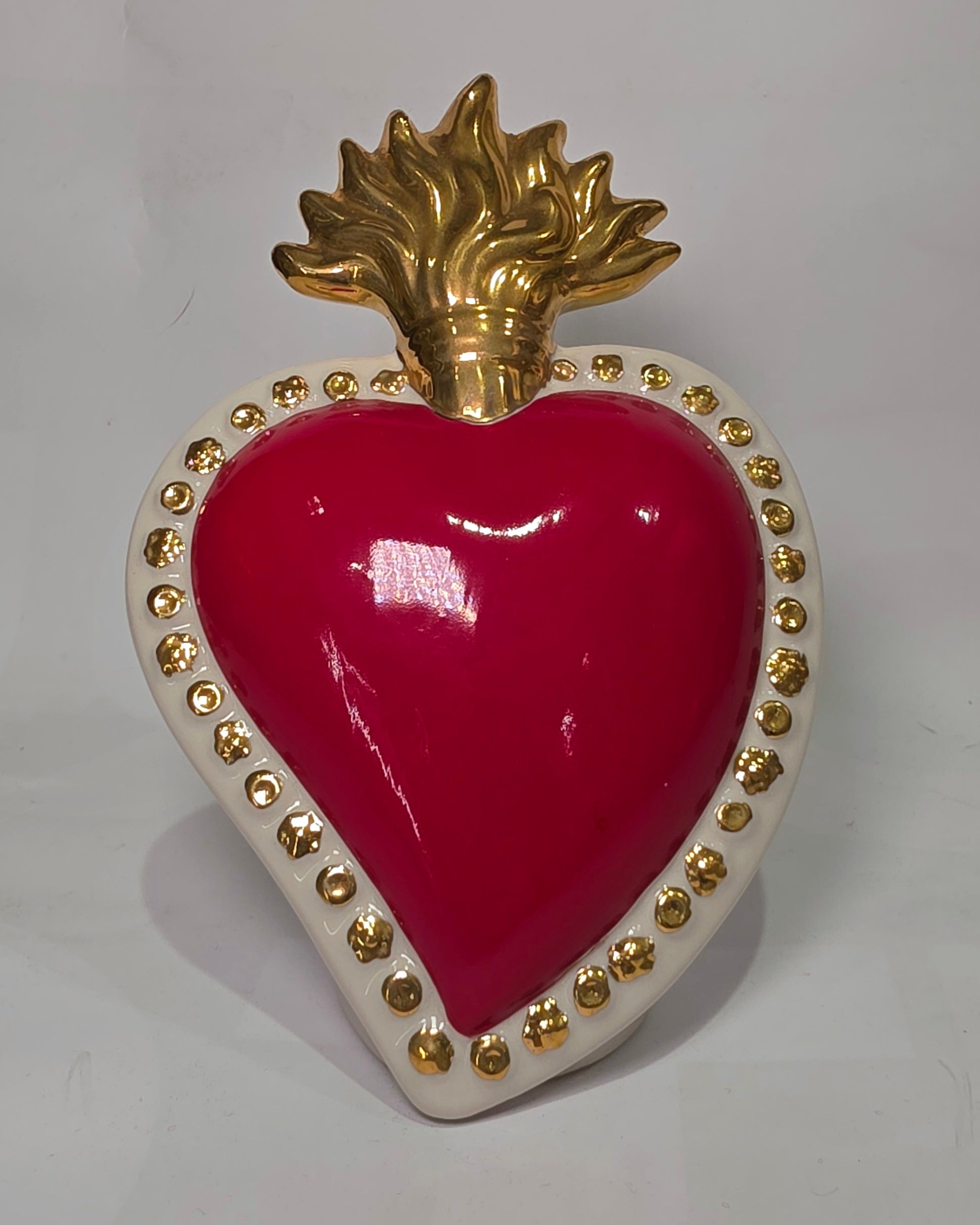 Scultura a forma di Cuore Sacro in ceramica rossa lucida, completata da una fiamma superiore dorata e una cornice bianca perlinata con borchie oro. Questo oggetto di design artigianale con base d'appoggio è un complemento d'arredo iconico, ideale per donare un tocco di eleganza e spiritualità moderna agli interni.