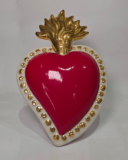 Scultura a forma di Cuore Sacro in ceramica rossa lucida, completata da una fiamma superiore dorata e una cornice bianca perlinata con borchie oro. Questo oggetto di design artigianale con base d'appoggio è un complemento d'arredo iconico, ideale per donare un tocco di eleganza e spiritualità moderna agli interni.