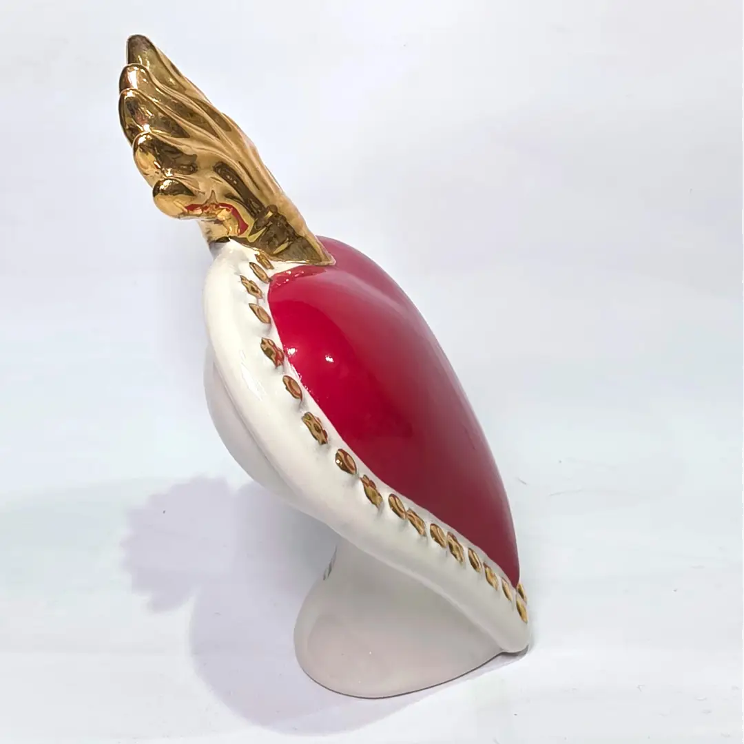 Scultura a forma di Cuore Sacro in ceramica rossa lucida, completata da una fiamma superiore dorata e una cornice bianca perlinata con borchie oro. Questo oggetto di design artigianale con base d'appoggio è un complemento d'arredo iconico, ideale per donare un tocco di eleganza e spiritualità moderna agli interni.
