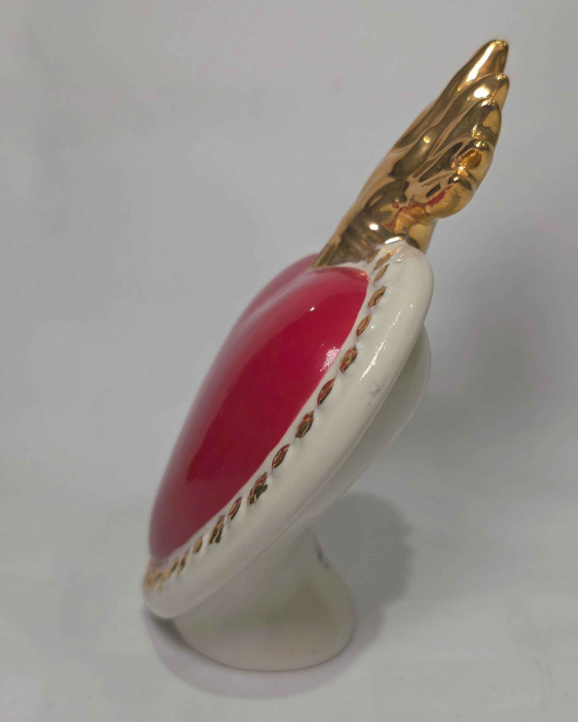 Scultura a forma di Cuore Sacro in ceramica rossa lucida, completata da una fiamma superiore dorata e una cornice bianca perlinata con borchie oro. Questo oggetto di design artigianale con base d'appoggio è un complemento d'arredo iconico, ideale per donare un tocco di eleganza e spiritualità moderna agli interni.