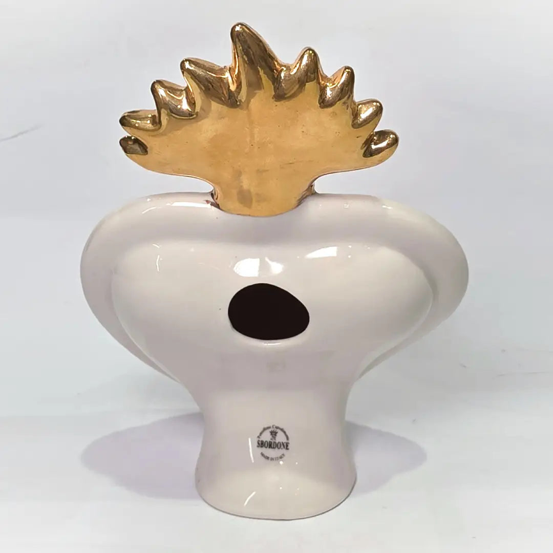 Scultura a forma di Cuore Sacro in ceramica rossa lucida, completata da una fiamma superiore dorata e una cornice bianca perlinata con borchie oro. Questo oggetto di design artigianale con base d'appoggio è un complemento d'arredo iconico, ideale per donare un tocco di eleganza e spiritualità moderna agli interni.