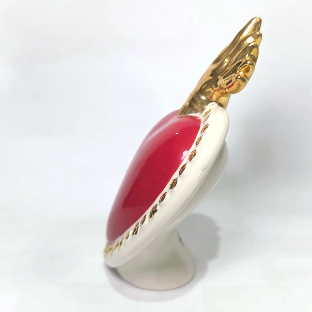 Scultura a forma di Cuore Sacro in ceramica rossa lucida, completata da una fiamma superiore dorata e una cornice bianca perlinata con borchie oro. Questo oggetto di design artigianale con base d'appoggio è un complemento d'arredo iconico, ideale per donare un tocco di eleganza e spiritualità moderna agli interni.