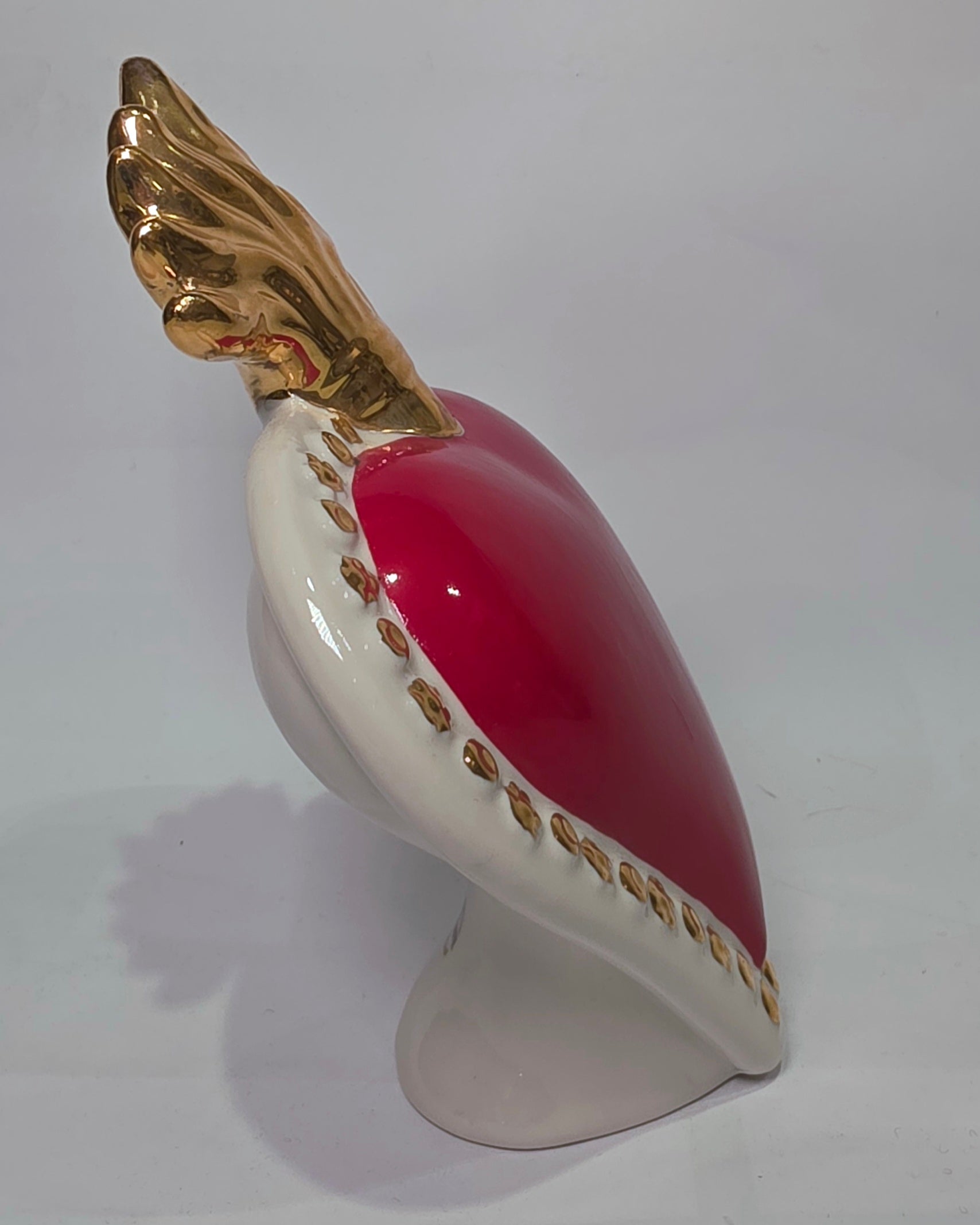 Scultura a forma di Cuore Sacro in ceramica rossa lucida, completata da una fiamma superiore dorata e una cornice bianca perlinata con borchie oro. Questo oggetto di design artigianale con base d'appoggio è un complemento d'arredo iconico, ideale per donare un tocco di eleganza e spiritualità moderna agli interni.